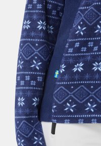 Granatowy polarowy pulower z białymi geometrycznymi wzorami i płatkami śniegu. Posiada boczny zamek błyskawiczny i małą metkę ze szwedzką flagą. Miękka faktura.