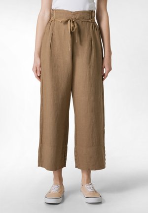 Pantaloni ampi in lino color marrone chiaro con vita elasticizzata e dettaglio a fiocco, dotati di tasche laterali e una finitura testurizzata. Abbinati a sneakers beige.