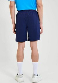 Persoon in donkerblauwe sportshorts, witte sokken, witte sneakers en een blauw shirt, staand met de rug naar de camera op een effen achtergrond.