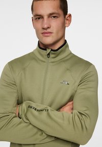 Zielona oliwkowa kurtka zapinana na zamek z wysokim kołnierzem, posiadająca czarne wykończenie wewnętrzne oraz nadrukowane logo na klatce piersiowej i rękawie. Gładka faktura materiału.