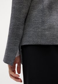 Gros plan d'une main à côté d'une manche de veste grise à motif chevrons et d'un pantalon noir sur fond blanc.