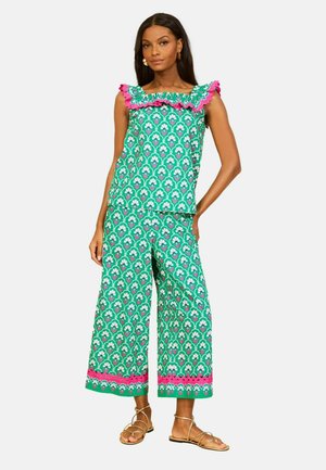 Femme portant un haut sans manches vert à motifs avec bordure à volants rose et pantalon large court assorti, associé à des sandales dorées à brides.