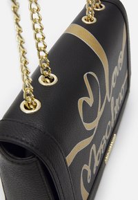 Borsa a tracolla in pelle nera con catene dorate, caratterizzata da una superficie liscia e un audace design con lettering dorato. Forma quadrata con lati strutturati.