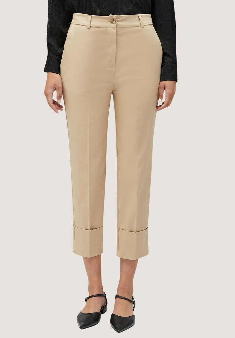 Pantalon beige sur mesure coupé court avec ourlets retroussés, porté avec des chaussures noires à bout pointu et bride à la cheville et un haut noir texturé à manches longues.