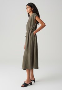 Olivgrünes, ärmelloses Maxikleid mit elastischem Taillenbund, seitlichen Taschen und fließender Textur. Kombiniert mit schwarzen Blockabsatzsandalen.