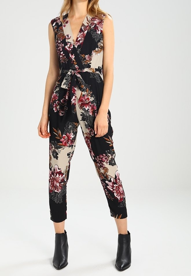 Combinaison florale avec un design enveloppant en noir, beige et rose, dotée d'une taille nouée, sans manches et avec des jambes raccourcies. Bottines noires.