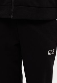 Pantaloni jogger neri in tessuto liscio, con vita con zip e un piccolo logo EA7 bianco sul lato sinistro.