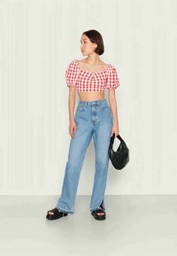 Mulher vestindo um crop top de mangas bufantes em xadrez vermelho e branco, jeans de cintura alta azul claro com fendas laterais, sandálias pretas, segurando uma bolsa preta.