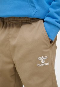 Bež Hummel hlače z elastičnim pasom, z vezenim logotipom na stegnu in roko v žepu, združene z modro majico z dolgimi rokavi.