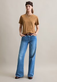 Braunes kurzärmliges T-Shirt, kombiniert mit hellblauen weitem Jeans. Das Outfit wird durch einen braunen Gürtel und ein gemustertes Haarband ergänzt.