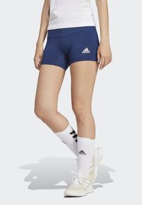 Pantaloni sportivi navy con vita elasticizzata, texture liscia e logo Adidas; abbinati a calze bianche fino al ginocchio e scarpe sportive.