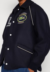 Námořnicky modrý kabát z vlněné směsi s žebrovaným límcem, dvěma bočními kapsami, slonovinovým lemováním a vyšívaným logem Lacoste na hrudi.