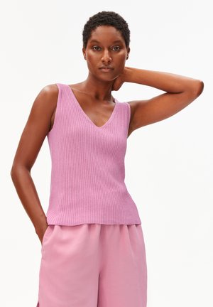 Vrouw met kort krullend haar, gekleed in een mouwloos roze geribd V-hals topje en losse roze broek, poserend met één hand achter haar nek.