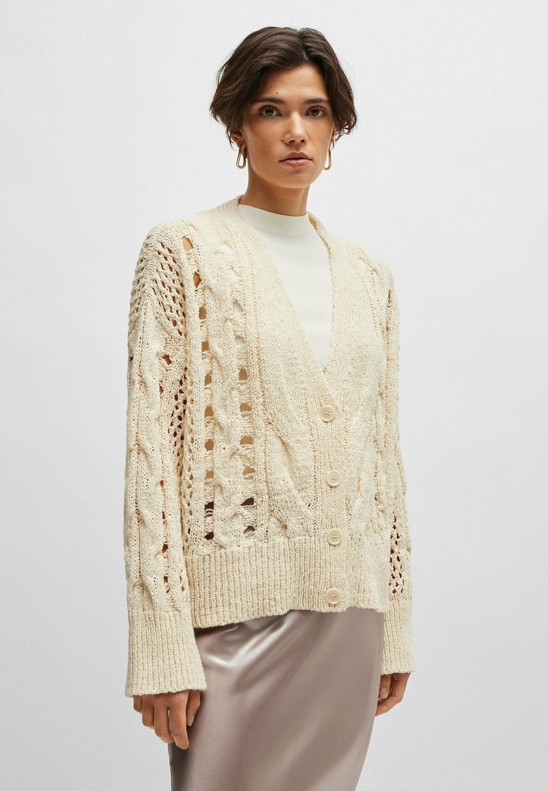 Cardigan a maglia crema con scollo a V profondo, caratterizzato da motivi a treccia, dettagli a traforo e cinque bottoni sulla parte anteriore.