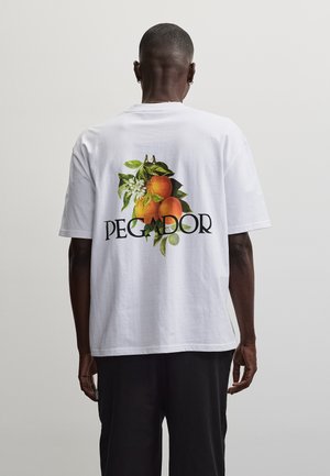 Bărbat purtând un tricou alb cu grafică de portocale, frunze și flori deasupra cuvântului „PEGADOR” pe spate, stând într-un fundal simplu.