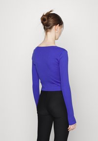 Długi rękaw, krótki top w intensywnym purpurowym prążkowanym materiale. Charakteryzuje się szerokim dekoltem i dopasowanym krojem, zestawiony z czarnymi legginsami.