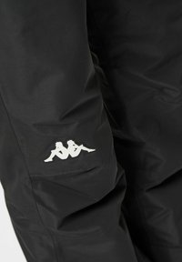 Tissu de pantalon noir montrant un petit logo blanc de deux figures assises en miroir dos à dos sur la partie supérieure de la cuisse.