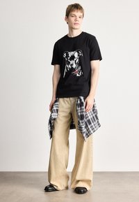 T-shirt de algodão preto com uma estampa gráfica de cachorro, calças largas bege e uma camisa cinza de xadrez atada à cintura. Sapatos pretos completam o visual.