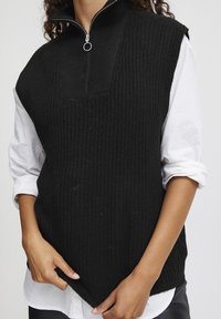 Schwarzer gestrickter ärmelloser Pullover mit gerippter Textur, hohem Kragen und silbernem Reißverschlussdetail. Über einem weißen Hemd getragen, was einen geschichteten Look zeigt.