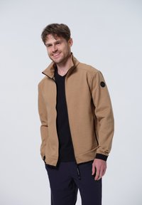 Chaqueta tipo sudadera de color marrón con cuello alto y puños acanalados, hecha de una tela suave. Presenta un parche con logo negro en el hombro izquierdo y bolsillos con cremallera.