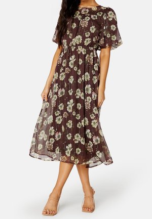 Vrouw die een bruine midi-jurk met pofmouwen en een wit bloemmotief draagt, gecombineerd met naakte open-toe hakken, staand met één been gekruist.