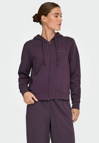 Violets ZIP-uppis džemperis, kas izgatavots no mīksta materiāla. Tam ir kapuce, priekšējās kabatas un minimāls zīmols uz krūškurvja.