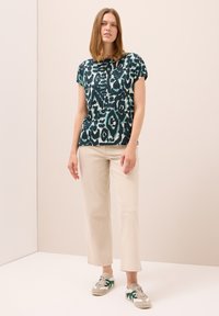 Mujer de pie con una blusa de estampado abstracto verde y negro de manga corta, pantalones rectos beige y zapatillas beige con acentos verdes.