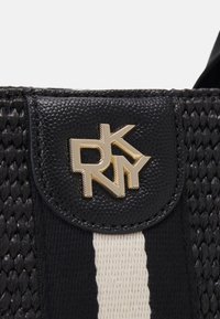 Gros plan d’un patch en cuir texturé noir avec le logo doré DKNY sur un fond de tissu tissé rayé noir et blanc.