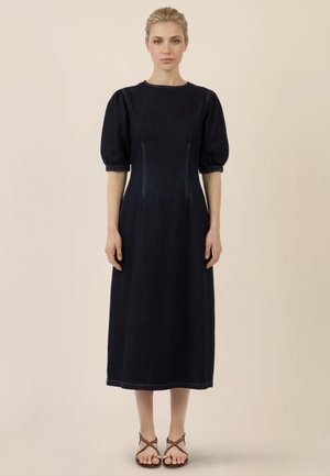 Frau steht und trägt ein mittellanges schwarzes Kleid mit Puffärmeln und braune Riemchensandalen vor beigem Hintergrund.