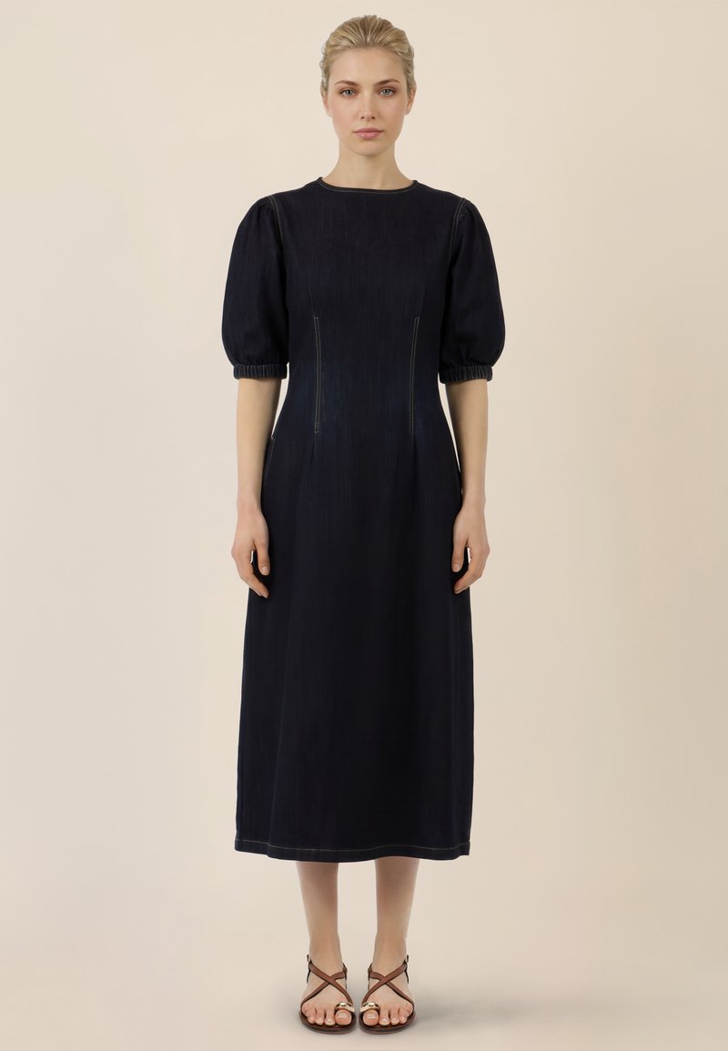 Femme debout portant une robe midi noire à manches bouffantes et des sandales à lanières marron sur fond beige.