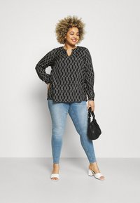 Schwarze langärmlige Bluse mit einem geometrischen weißen Muster, einem Schlüssellochausschnitt, kombiniert mit hellblauen Skinny-Jeans und weißen Blockabsatzsandalen.