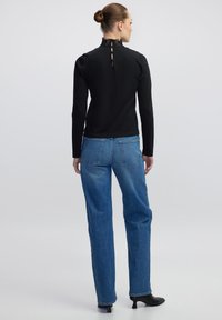 Zwarte langemouwtop met kantel_detail, gecombineerd met mid-rise blauwe denim jeans. Model draagt zwarte hakken.