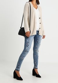 Béžový pletený cardigan s knoflíky, bílý top, modré džíny zdobené květinovými vzory a černé lodičky na vysokém podpatku.