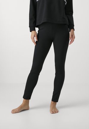 Leggings neri realizzati in tessuto elastico, design aderente e gambe affusolate, abbinati a una felpa nera e piedi nudi.