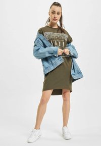 Oliwkowa sukienka t-shirtowa z białym nadrukiem graficznym, zestawiona z jasnoniebieską dżinsową kurtką. Białe sneakersy dopełniają cały strój.