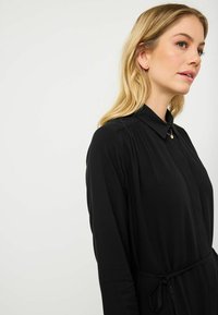 Czarna sukienka z długimi rękawami i kołnierzykiem, z zapięciem na guziki i paskiem w talii. Gładka tekstura i minimalistyczny design.