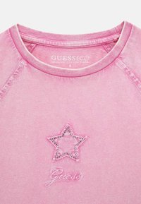 Rožinė organinės medvilnės marškinėliai su žvaigždės dizainu, išsiuvinėtu sidabriniu kontūru, ir prekės ženklo pavadinimu "Guess" žodiniu šriftu.