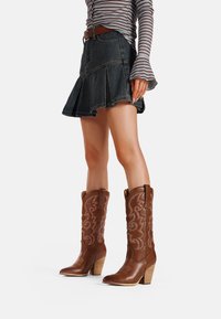 Stivali da cowboy in pelle marrone con cuciture intricate, punta affilata e tacco a cascata, abbinati a una gonna in denim plissettata e una maglietta a righe.