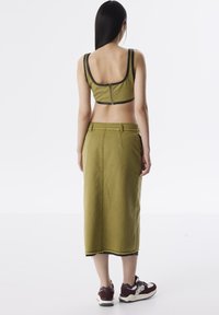 Top court vert olive avec des bordures noires et des détails de fermeture éclair, associé à une jupe longue assortie. Porté avec des baskets rouge foncé et crème.