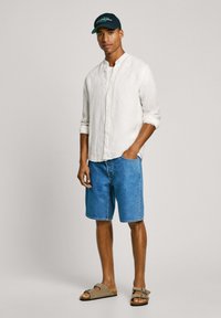 Camicia in lino leggero di colore avorio, pantaloni shorts in denim blu, sandali marroni e un cappellino scuro con un logo, in piedi su uno sfondo neutro.
