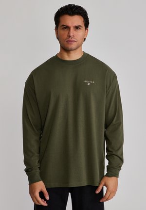 Mand, der vender fremad, iført en olivengrøn langærmet skjorte med "SikSilk"-logo på venstre bryst, stående foran en ensfarvet baggrund.