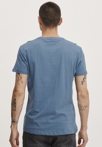 Blå kortærmet bomulds T-shirt med rund halsudskæring, glat tekstur og tætsiddende silhuet, vist fra ryggen med tatoveringer synlige på armene.