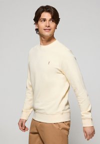 Sudadera amarilla clara con cuello redondo, mangas largas y puños de canalé. Presenta un pequeño logo en contraste en el pecho. Textura suave.