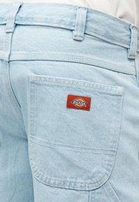 Τσέπη από ανοιχτό μπλε τζιν με μια ραμμένη ετικέτα Dickies κόκκινου και κίτρινου χρώματος πάνω από την τσέπη.