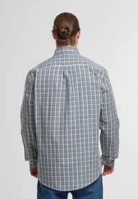 Mann mit einem Dutt, der ein langärmliges, blau-weiß kariertes Hemd trägt und mit dem Rücken zur Kamera vor einem schlichten Hintergrund steht.