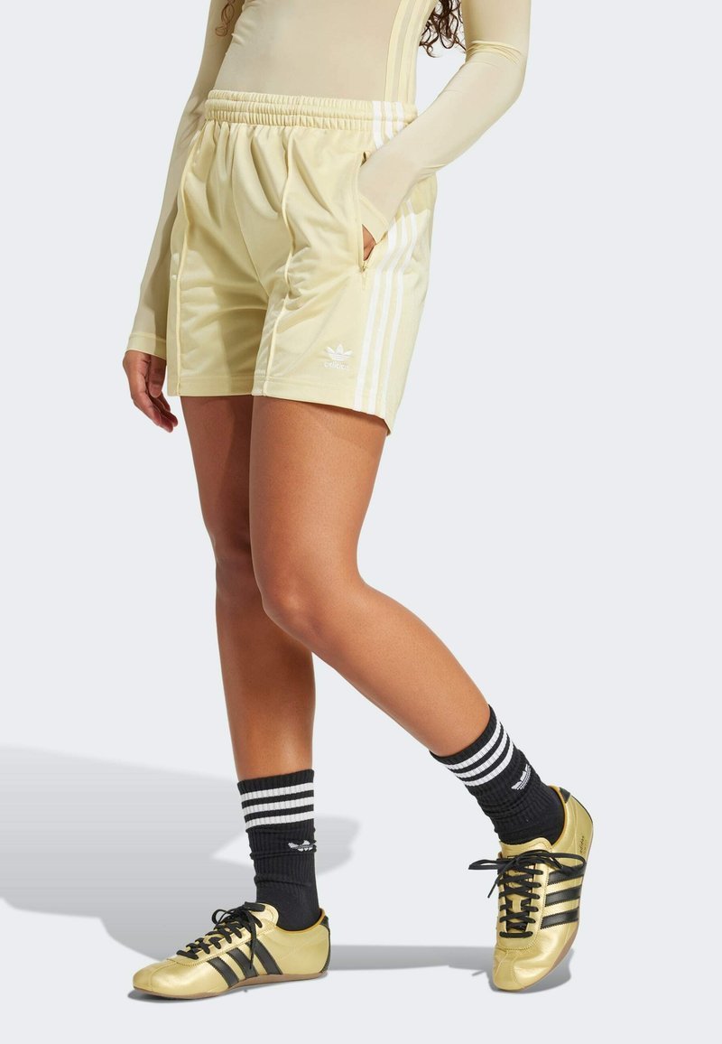 Lichtgele sportshorts met een elastische tailleband, twee zijzakken en drie witte strepen aan elke zijde, gecombineerd met zwarte en gouden schoenen.