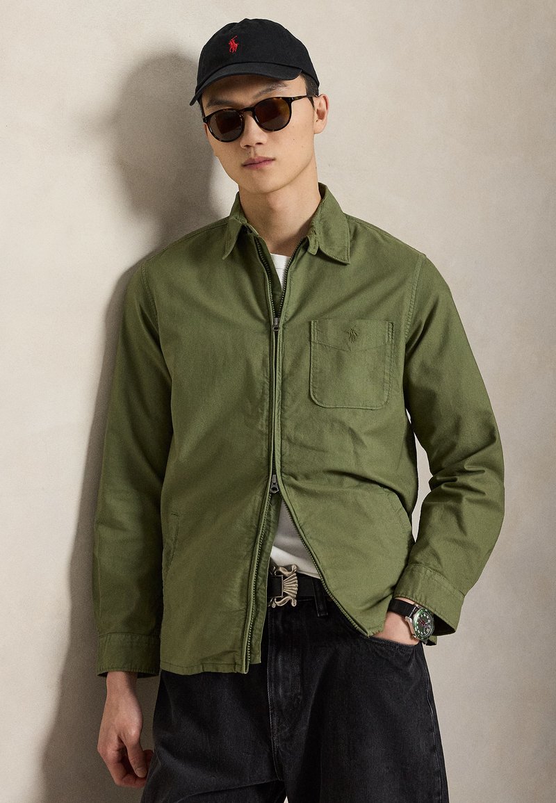 Junger Mann trägt olivgrüne Jacke mit Reißverschluss, schwarze Kappe mit rotem Logo, dunkle Sonnenbrille, schwarze Jeans und Armbanduhr, steht vor neutraler Wand.