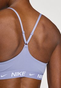 Μωβ αθλητικό σουτιέν με λεπτές τιράντες, ρυθμιζόμενο εξάρτημα και φαρδιά ζώνη με το λογότυπο της Nike σε λευκό. Ομαλή υφή υφάσματος.