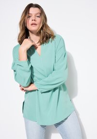 Blusa verde menta con scollo a V, maniche arricciate e fessure laterali. Abbinata a jeans azzurri chiari, in tessuto morbido e vestibilità comoda.