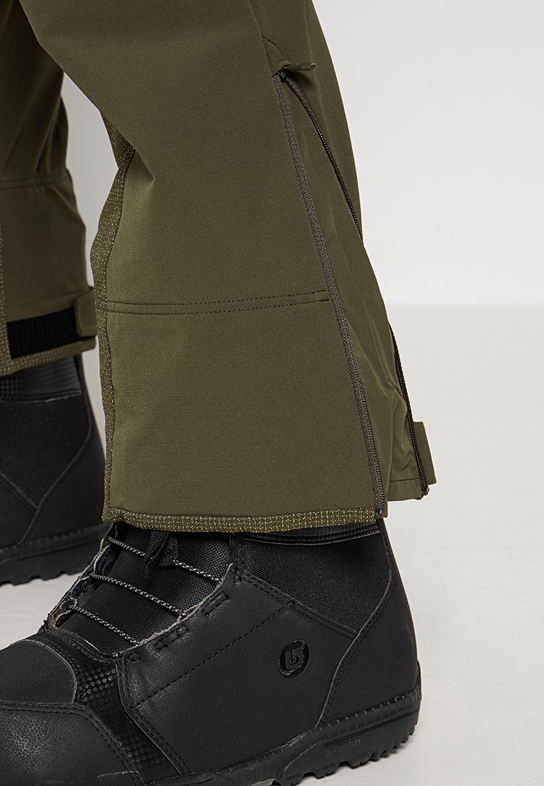 Olivgrüne wasserdichte Hosen mit Reißverschlüssen an den Knöcheln, kombiniert mit schwarzen Wanderschuhen mit strukturierten Oberflächen und Schnürsenkeln.